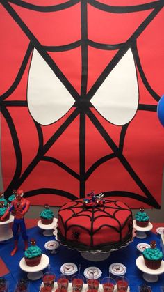 a spiderman themed birthday cake and cupcakes on a blue tablecloth in front of a red backdrop