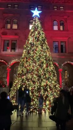 a large christmas tree is lit up in front of a building with people standing around it