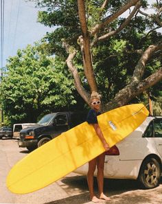 a woman standing next to a tree holding a yellow surfboard in front of her