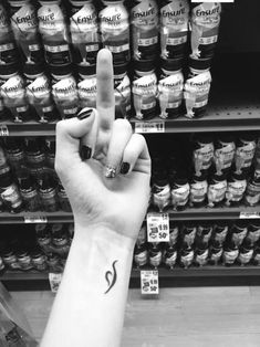 a woman's hand with a small tattoo on the middle finger, in front of shelves of condiments