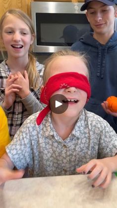 three children with blindfolds are sitting at a table and one child is holding an orange