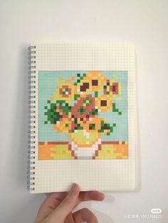 a hand is holding a notebook with a cross stitch pattern on it and there are flowers in the vase