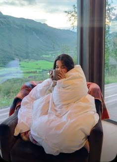 a woman wrapped in a blanket is sitting on a chair and looking out the window