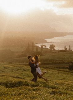 a man and woman standing on top of a lush green hillside next to the ocean