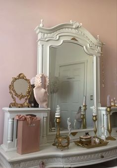 a white dressing table topped with a mirror and lots of gold accessories next to a pink wall