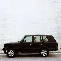 a black suv parked in front of a white brick wall with two people sitting in the driver's seat