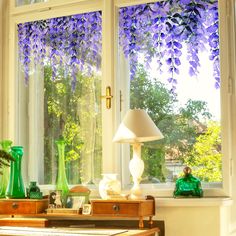 purple flowers are hanging over the windows in this living room with green glass vases