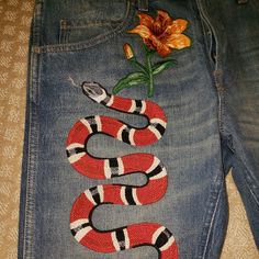 a pair of jeans with a snake embroidered on the front and side, sitting next to a flower