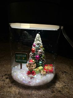 a small christmas tree in a glass jar filled with snow and other holiday decorations on top of a granite counter