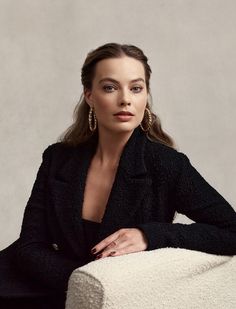 a woman sitting on top of a white chair wearing a black coat and gold earrings