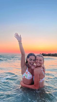 two women in bikinis sitting on the back of a boat at sunset, with their arms around each other