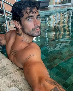 a man with tattoos sitting in a swimming pool next to a railing and looking off into the distance