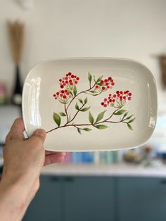 a hand holding a white plate with red berries on it and green leaves painted on the side