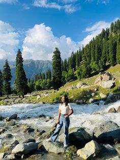 a woman standing on rocks in the middle of a river with mountains in the background