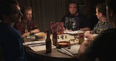 four men are sitting around a table with drinks and papers on it, looking at each other