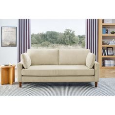 a beige couch sitting in front of a window next to a wooden table and bookshelf