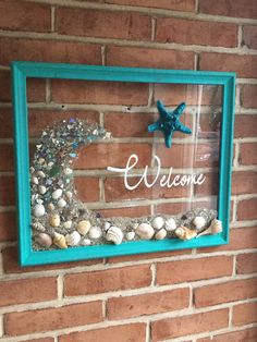 a welcome sign made out of seashells in a frame on a brick wall