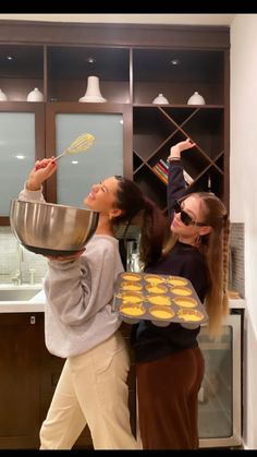 two young women are cooking in the kitchen with one holding a metal pan and another holding a spatula