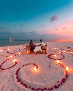 two people sitting at a table on the beach with candles in the shape of a heart