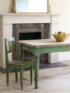 a green table with two chairs and a bowl of fruit on it in front of a fireplace