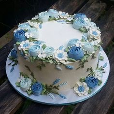 a white and blue cake with flowers on it sitting on a wooden table next to a bench