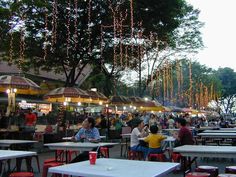 many people are sitting at tables in an outdoor restaurant area with lights strung from the trees