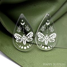 two clear earrings with white designs on them sitting on a green leather surface next to a purse