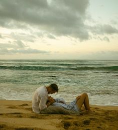 a man and woman sitting on top of a sandy beach next to the ocean under a cloudy sky