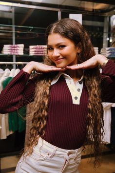 a woman with long curly hair standing in front of a rack of clothes and posing for the camera