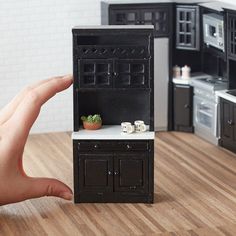 a hand is holding a miniature black kitchen with white counter tops and cabinets in the background