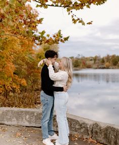 a man and woman standing next to each other near a body of water with fall leaves on the ground