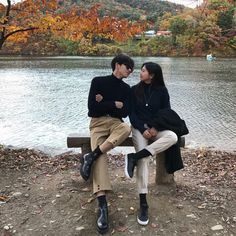 two people sitting on a bench next to a lake and trees with fall colors in the background