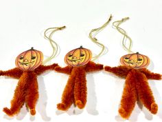 three stuffed animals with pumpkin faces hanging from strings on a white background, one is orange and the other is brown