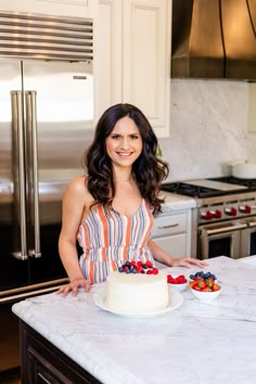 a woman standing in front of a cake on top of a kitchen counter next to a refrigerator