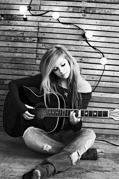 a woman sitting on the floor playing an acoustic guitar in front of a wooden wall