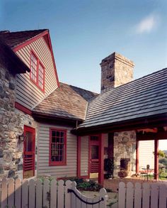 a stone house with red trim on the windows and shutters is shown in front of a white picket fence