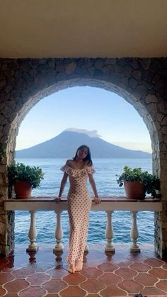 a woman in a dress standing on a balcony near the ocean with mountains in the background