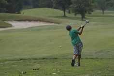 a man swinging a golf club at a ball on a green field with trees in the background