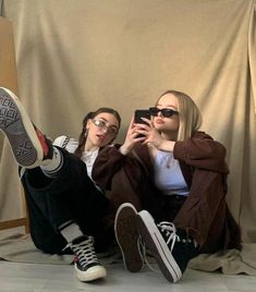 two young women sitting on the floor taking pictures with their cell phones, one is wearing sunglasses and the other has her legs crossed