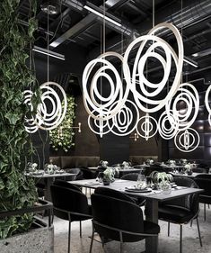 a dining room with black chairs and white chandeliers hanging from the ceiling, surrounded by greenery