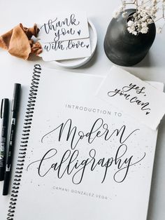 an open notebook with calligraphy on it next to two pens and a bowl of flowers