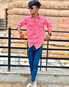 a man in pink shirt and blue jeans standing by a fence with his hands on his hips