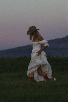 a woman in a white dress and hat is walking through the grass at dusk with her back to the camera