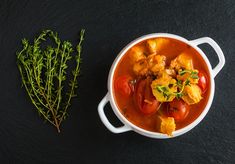 a bowl of stew next to a sprig of fresh herbs on a black surface