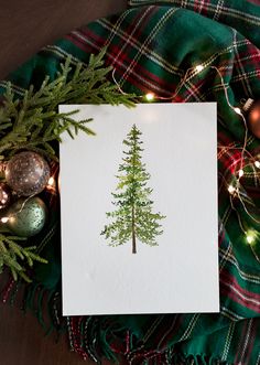 a card with a christmas tree drawn on it next to ornaments and a plaid blanket