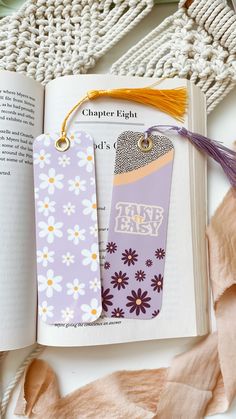 two bookmarks with tassels on them sitting next to an open book and scarf