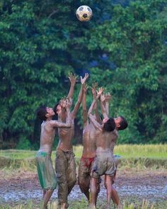 three boys are playing with a soccer ball in the mud while another boy jumps up to catch it