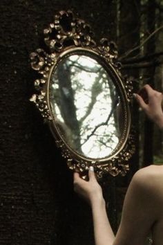 a woman holding a mirror in front of her face and looking at it through the trees