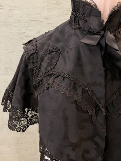 1800 vintage, half cape in black silk brocade with black beads designs trimmed in black lace. The lace around the bottom hem is wider and weightier than the lace at the high collar and around the beadwork at the shoulders. The cape is lined in silk. There is a black silk ribbon tie at the front. 2" stand up  collar, 16" from shoulder to shoulder in the back, 15" long, 3" lace trim at hem. Dry clean only Black Gothic Victorian Dress With Lace Trim, Black Victorian Dress With Lace Trim For Evening, Formal Black Capelet, Black Capelet For Costume, Elegant Black Capelet With Cape Sleeves, Black Victorian Dress With Lace Trim For Costume, Formal Black Victorian Dress With Lace Trim, Fitted Black Capelet, Black Fitted Capelet