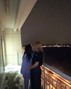 two people standing next to each other on a balcony at night with city lights in the background